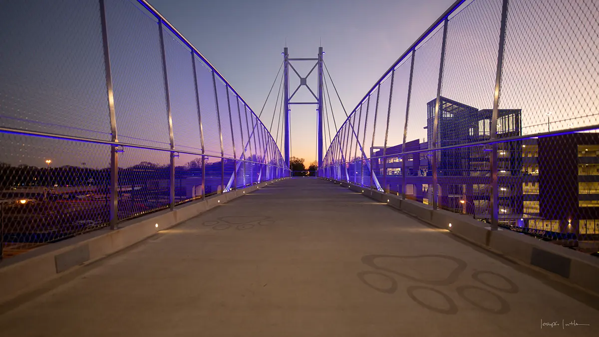 Illuminated pedestrian bridge under a twilight sky, with blue lights lining the walkway. City buildings are visible on the right, creating a serene urban scene.
