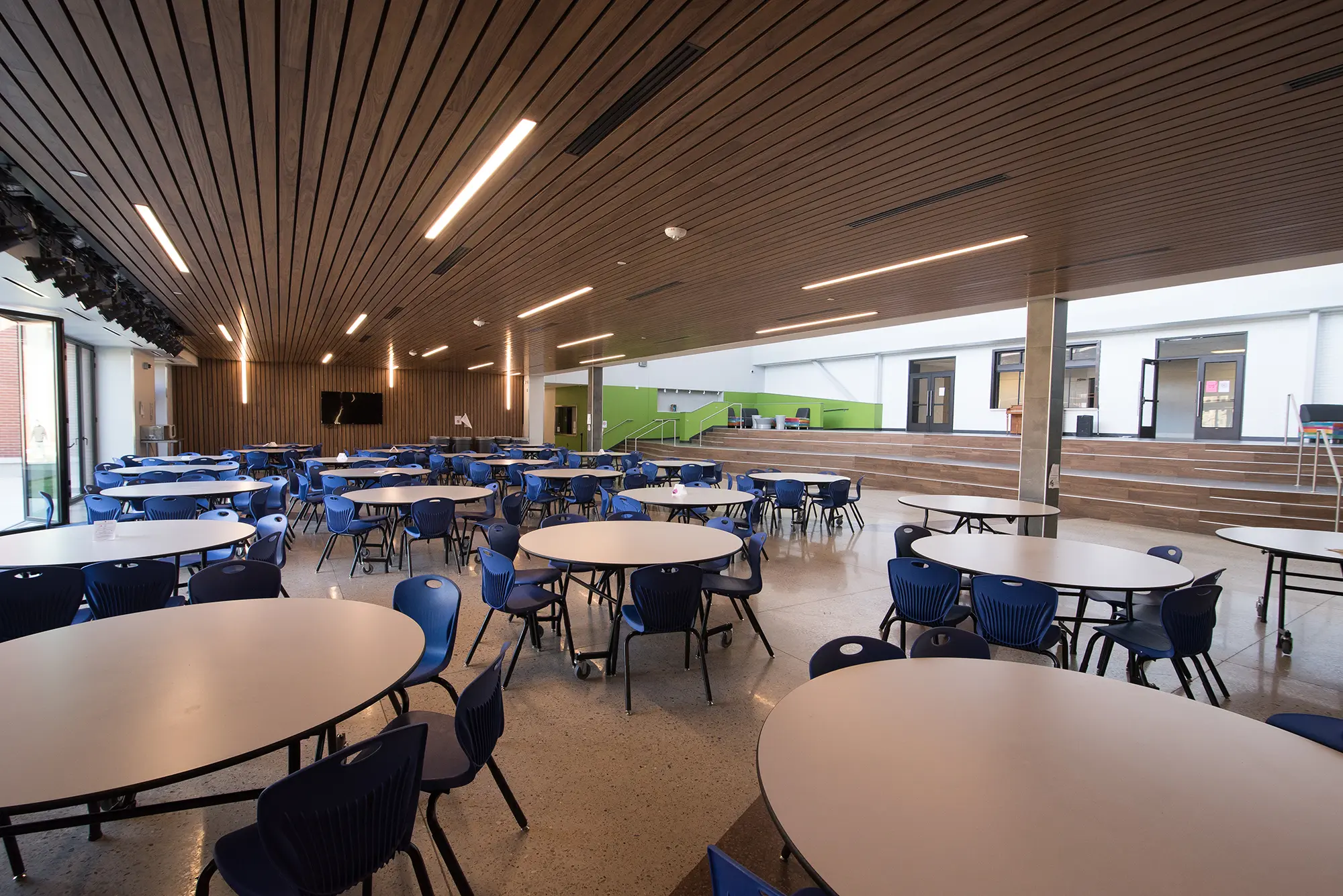 Spacious cafeteria with round tables and blue chairs on a polished floor, under a wooden ceiling with bright lights; airy and modern atmosphere.