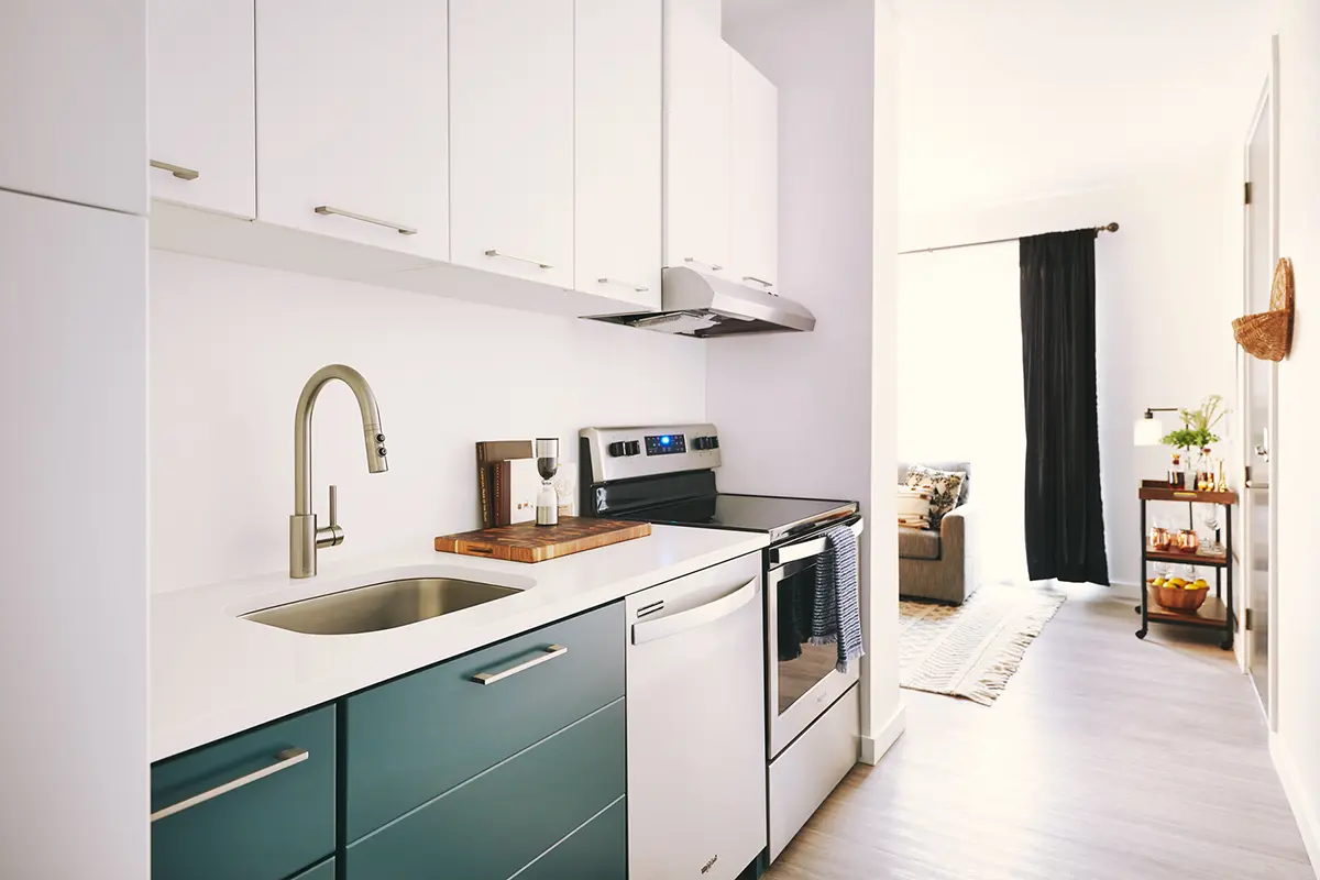 Cocina moderna con armarios blancos, cajones inferiores de color verde azulado y electrodomésticos de acero inoxidable. Espacio luminoso y minimalista que se abre a una sala de estar con luz natural.