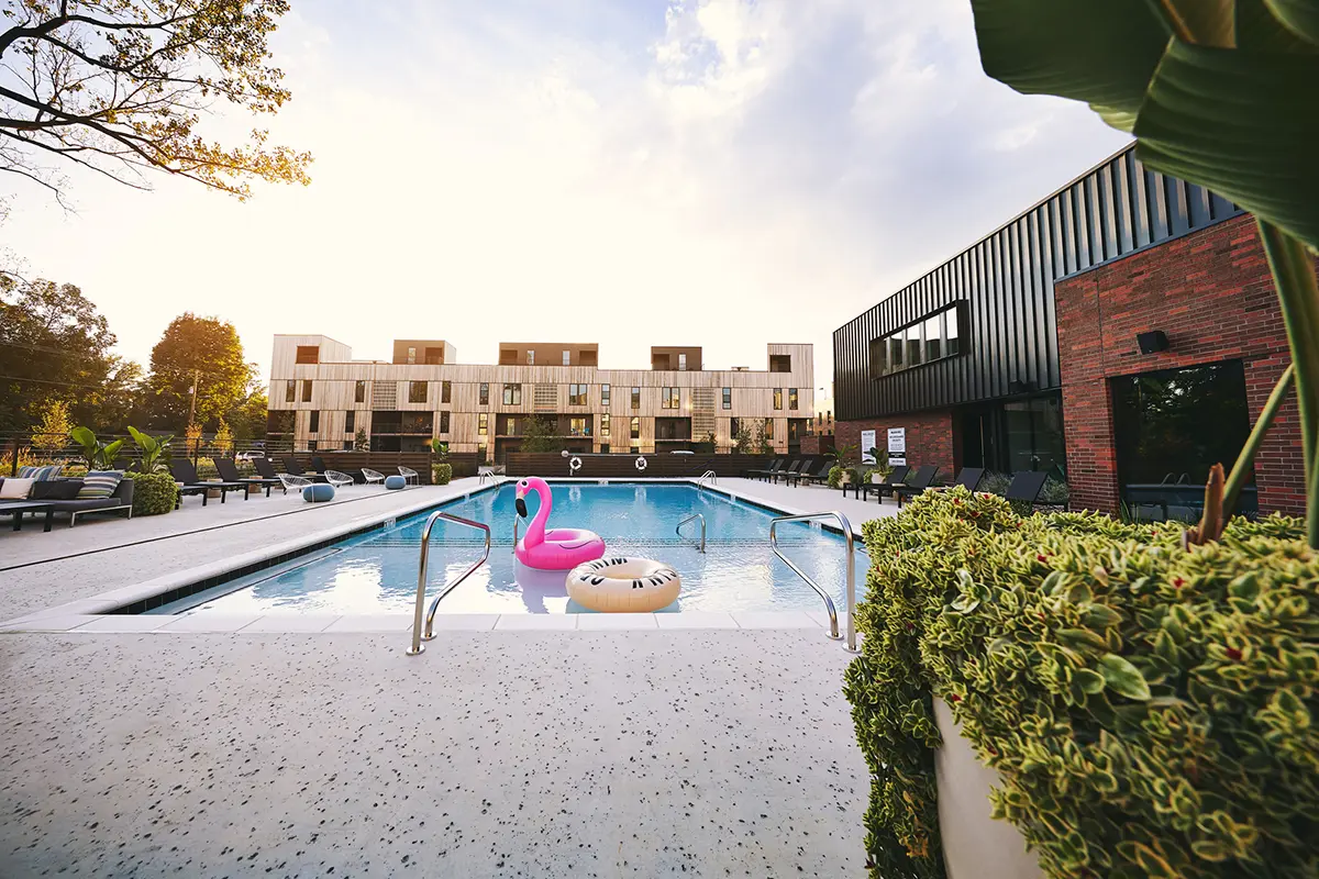 Piscina al aire libre con flotadores en forma de flamencos rosas y donuts, rodeada de modernos apartamentos y exuberante vegetación. El cielo está despejado y parcialmente nublado, lo que crea un ambiente relajado y acogedor.
