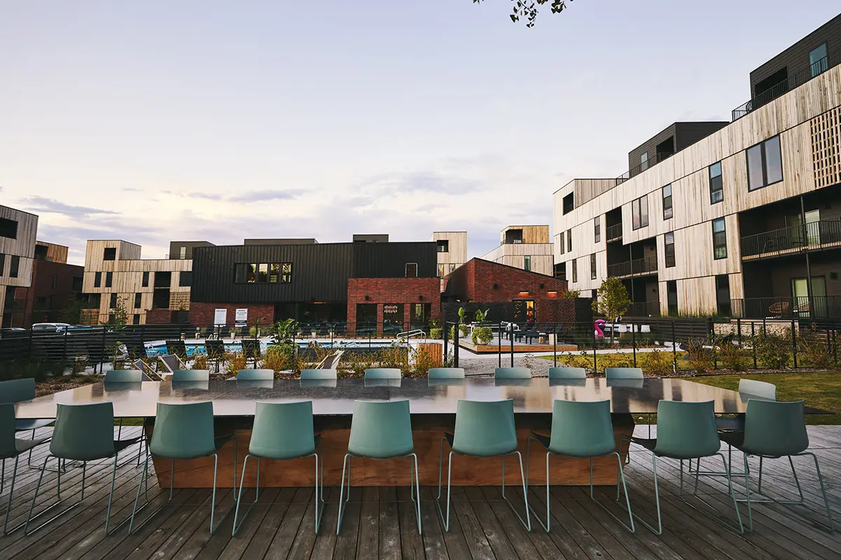 Ambiente moderno al aire libre con una mesa larga y sillas de color verde azulado en una terraza de madera. Alrededor hay edificios contemporáneos con grandes ventanas, bajo un cielo crepuscular.