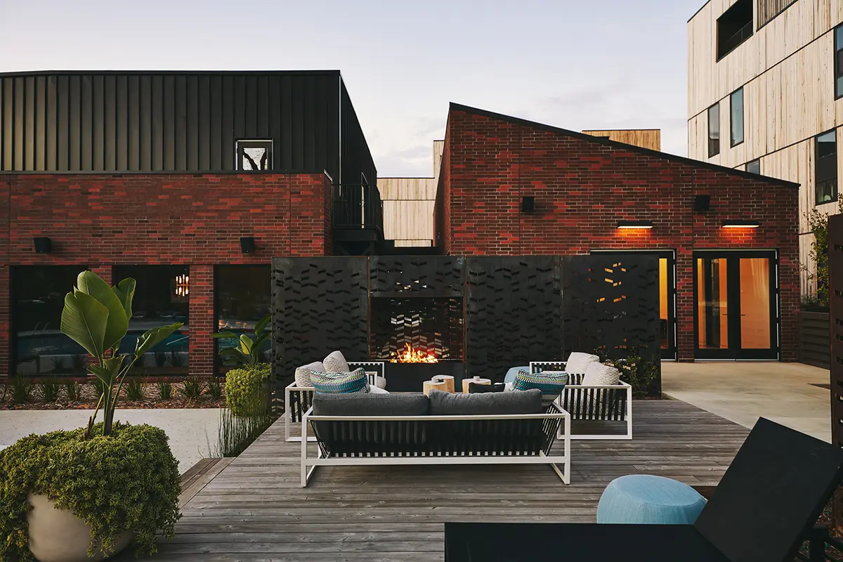 Patio exterior con mobiliario moderno y una chimenea encendida con los edificios de ladrillo como telón de fondo al atardecer; un ambiente acogedor y atractivo.