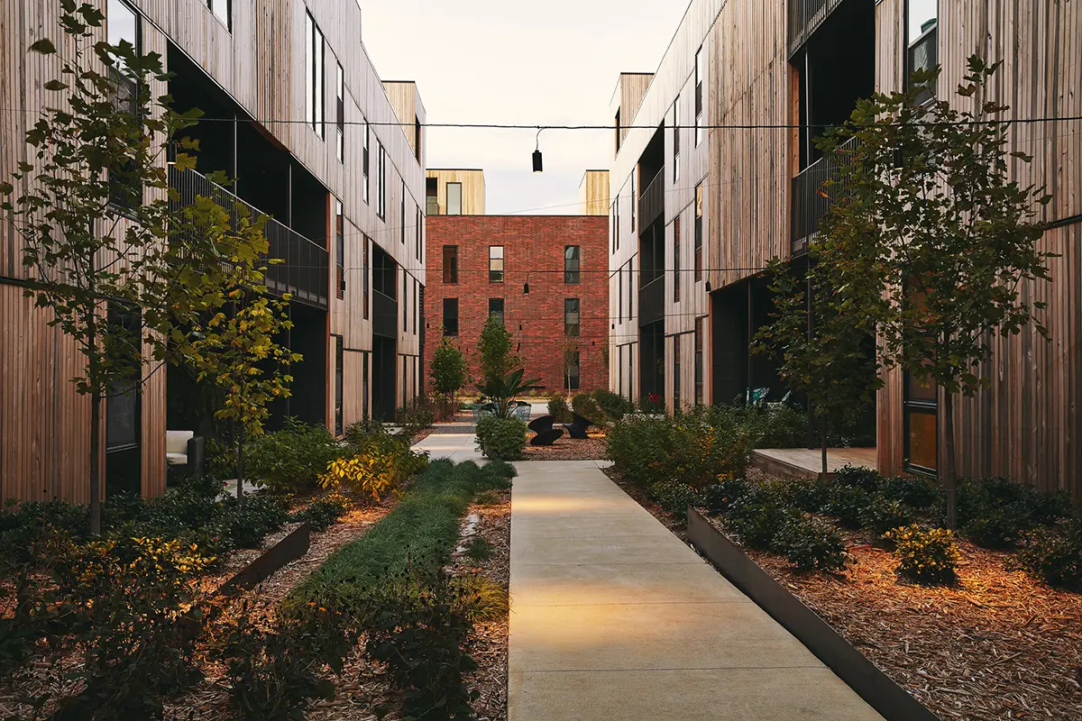 Patio moderno con fachadas de madera y un camino central, flanqueado por vegetación. Al fondo se ve un edificio de ladrillo rojo, bajo una suave iluminación.