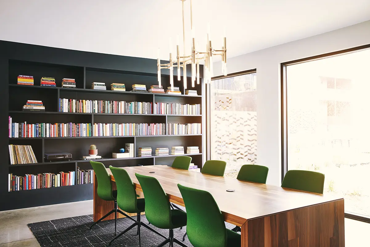 Una moderna sala de conferencias con una mesa de madera y seis sillas verdes. Sobre ella cuelga una elegante lámpara de araña. Una de las paredes cuenta con una gran estantería llena de libros, y una gran ventana permite la entrada de luz natural, creando un ambiente luminoso y acogedor.