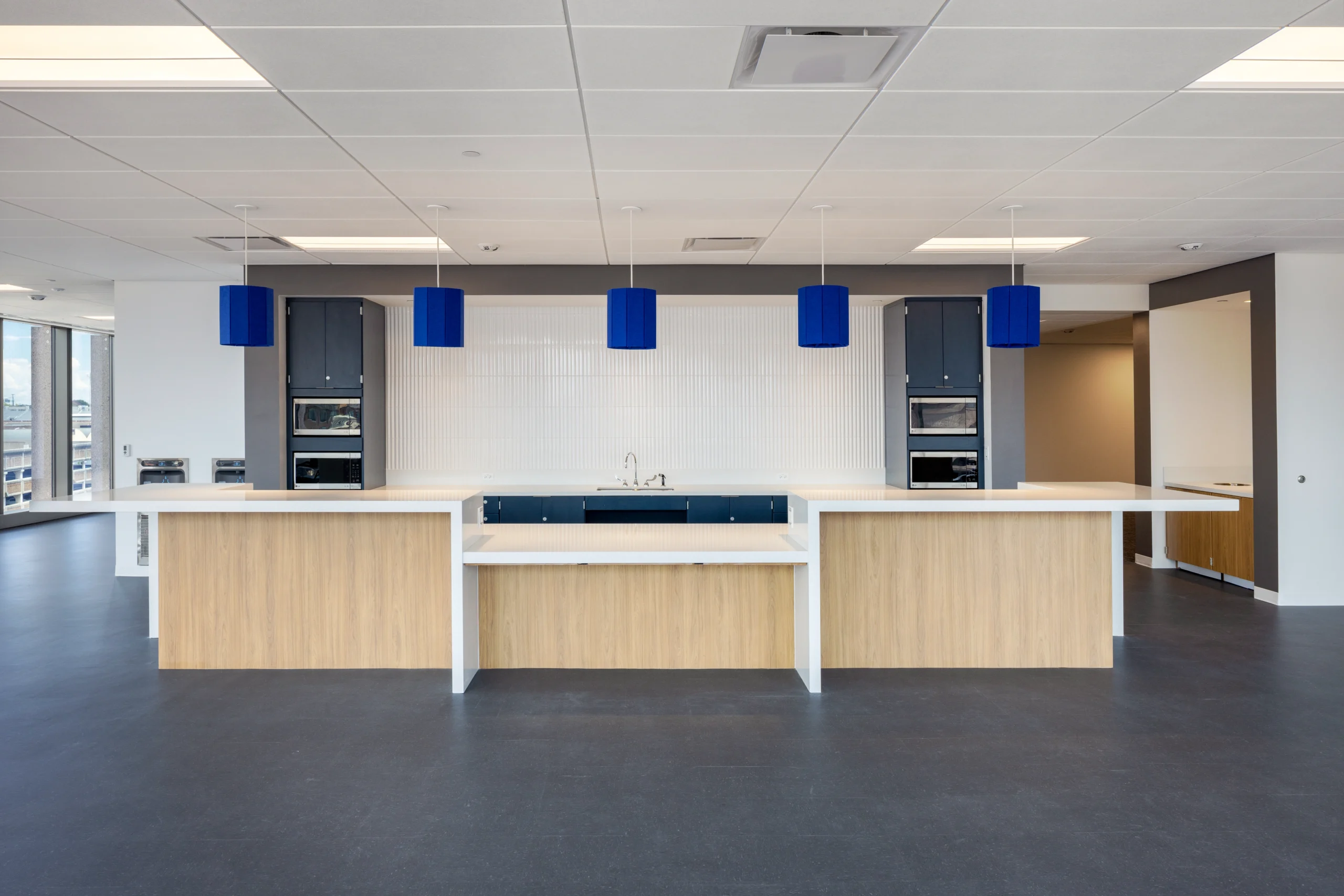 Modern kitchen with a minimalist design, featuring wood accents, blue pendant lights, a sleek counter, and built-in ovens against a white backsplash.