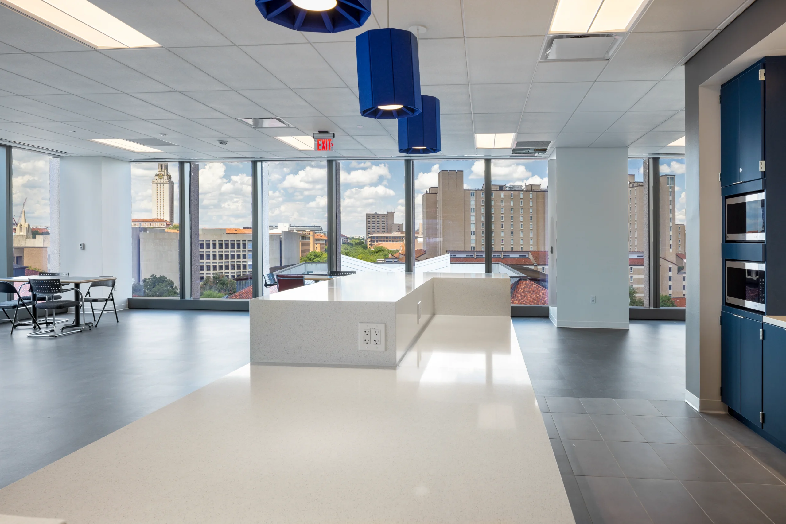 Modern office kitchen with blue pendant lights and white countertops. Large windows offer a cityscape view, creating an open, airy atmosphere.