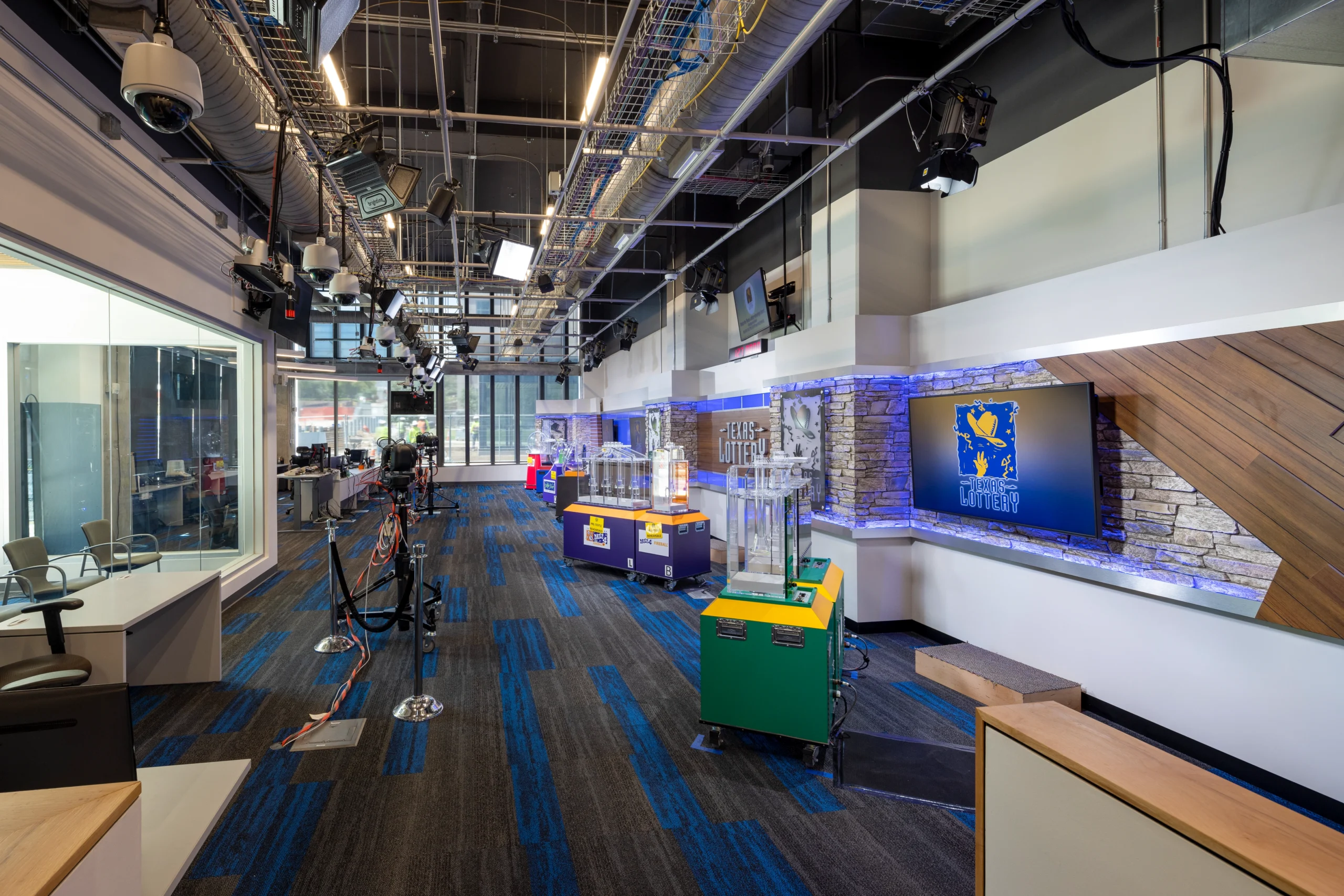 A brightly lit studio with lottery draw machines on a blue and gray carpet, modern lighting fixtures, and monitors displaying "Texas Lottery" logos on a stone wall.