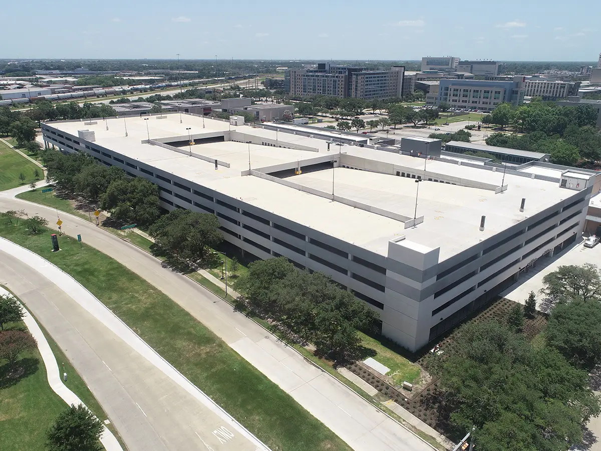 Aerial view of a large, multi-level parking garage in an urban area, surrounded by trees and roads. Nearby buildings and a clear sky create a calm atmosphere.