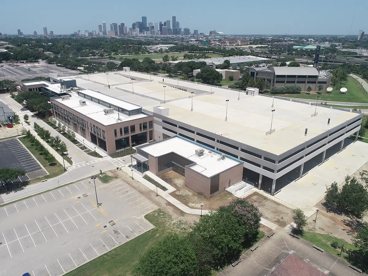Aerial view of a large, multi-level parking structure and adjacent low-rise building. Surrounding area includes parking lots, trees, and distant city skyline. Bright, clear day.