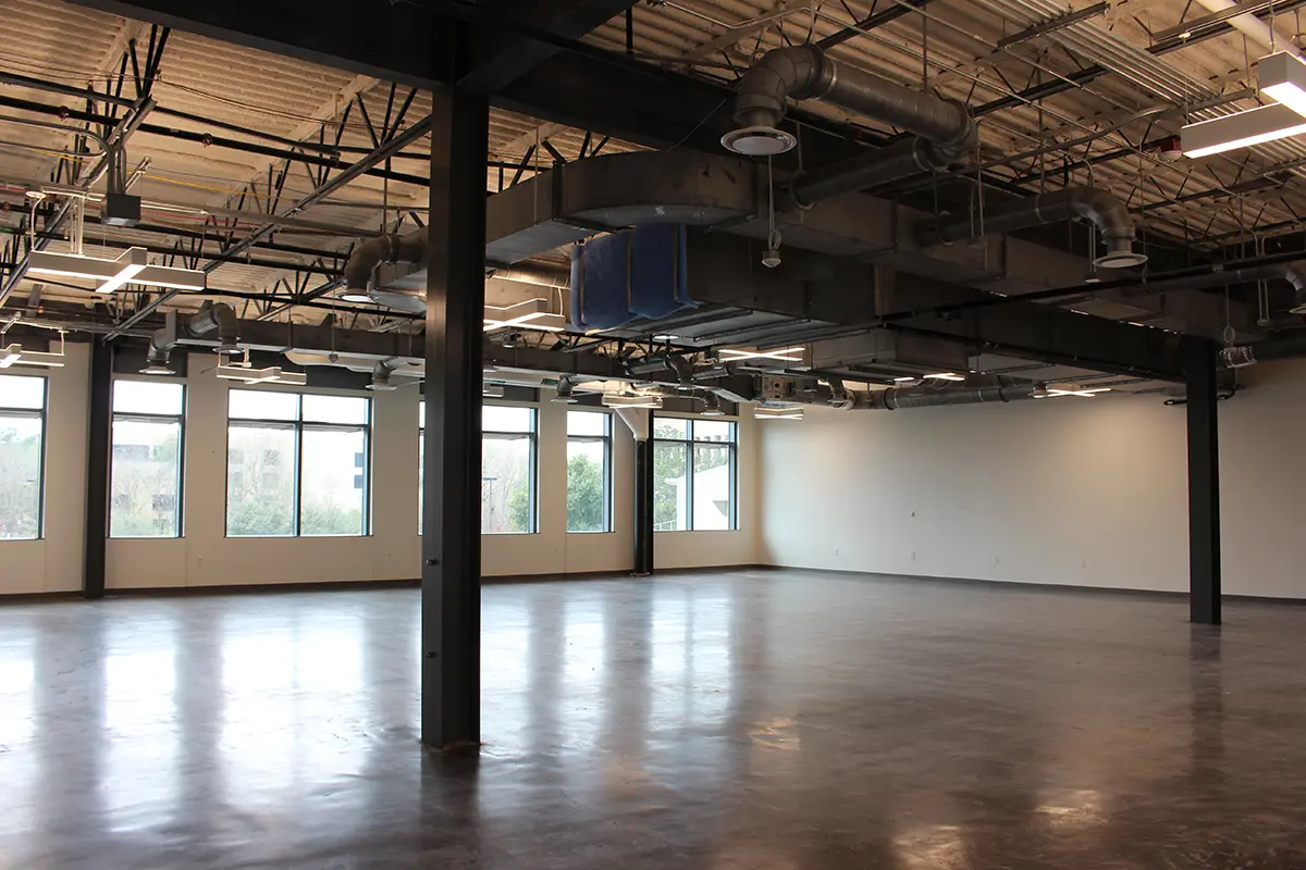Spacious, empty industrial loft with high ceilings, exposed ducts, large windows, and polished concrete floors. Bright natural light creates an open, airy feel.