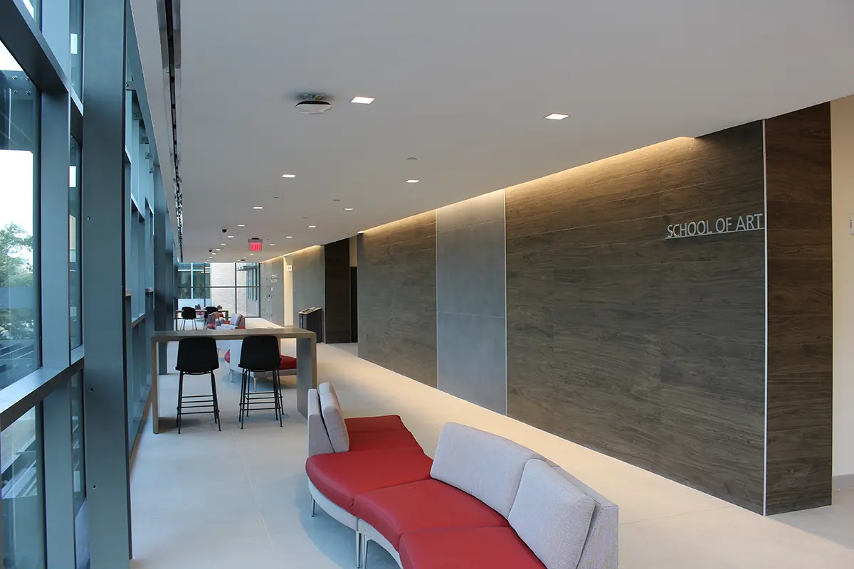 Modern hallway with large windows and sleek design. Red and gray seating curves along the wall; wooden panel labeled "School of Art." Bright, open atmosphere.