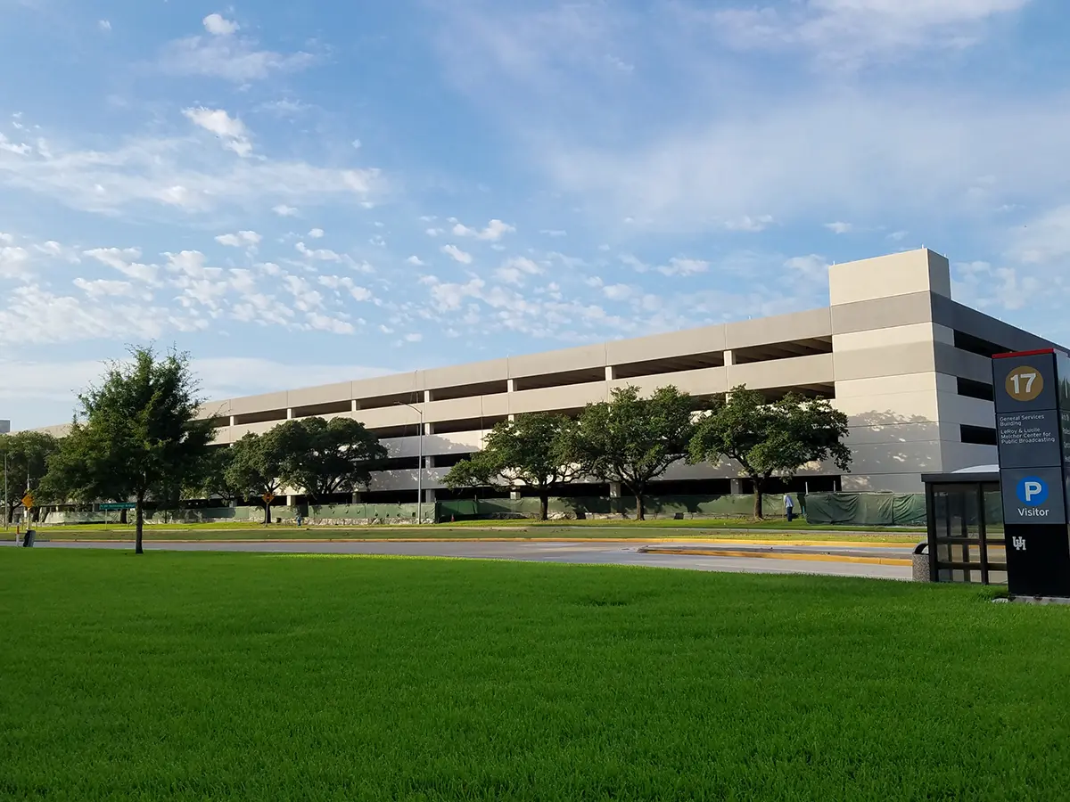 A five-story parking garage is surrounded by lush green grass and trees under a bright blue sky. A visitor parking sign is visible in the foreground.