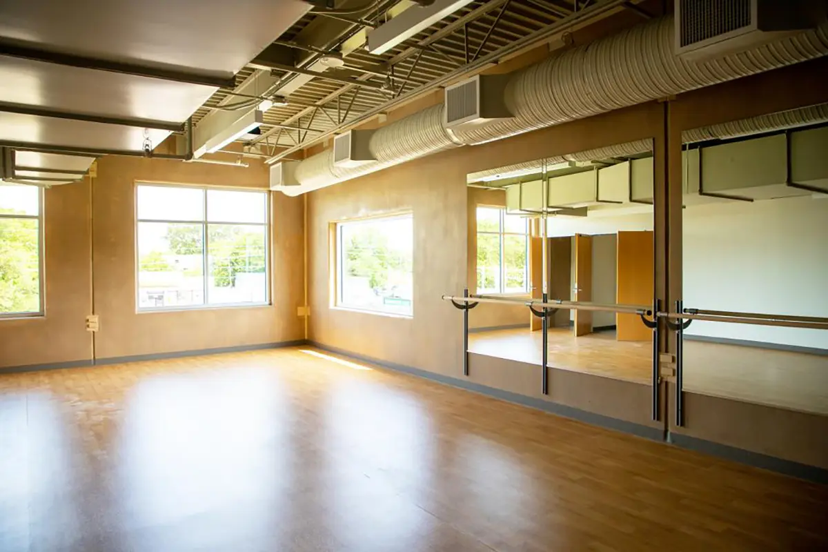 Spacious dance studio with wooden floors, mirrored wall, ballet barre, large windows, and exposed ductwork, creating a bright and open atmosphere.