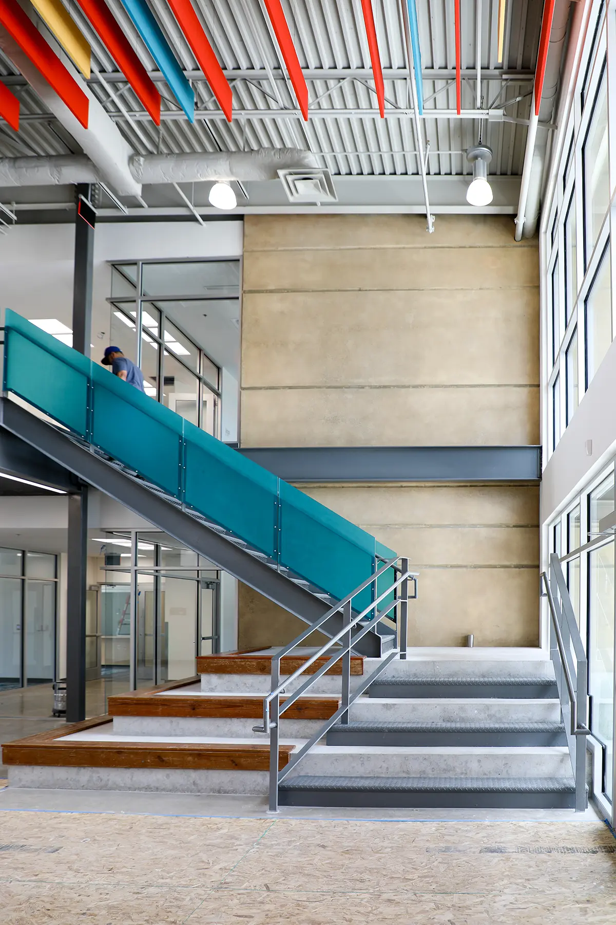 Modern interior with a teal glass-panel staircase, metal railings, and colorful ceiling beams. Natural light streams through large windows, creating a sleek, vibrant atmosphere.