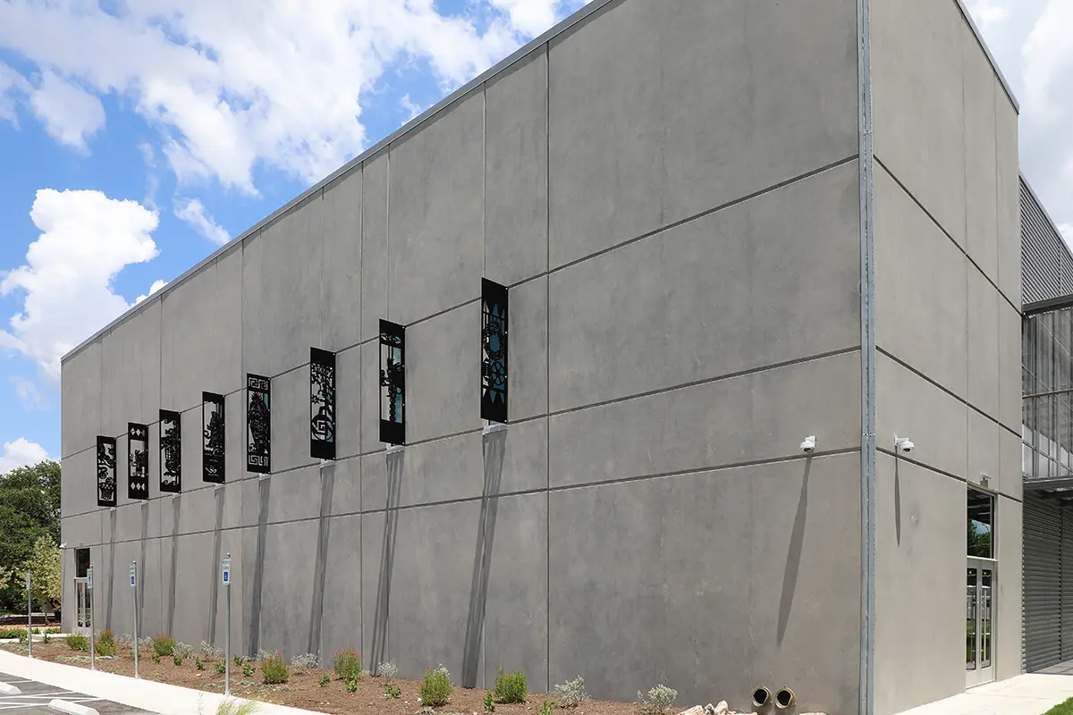 A modern gray concrete building with vertical rectangular windows featuring artistic lattice designs. The sky is blue with scattered clouds, creating a calm atmosphere.