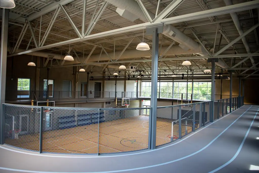 Indoor gym with a basketball court and overhead running track. The space is well-lit with large windows and metal beams, creating an open, airy feel.