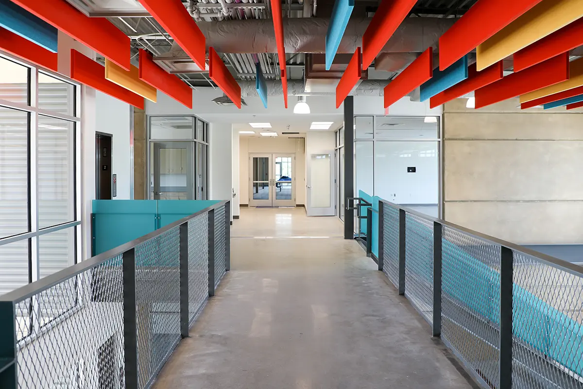 A modern hallway with colorful orange, blue, and yellow panels on the ceiling. Large windows and glass doors create an open, airy feel.