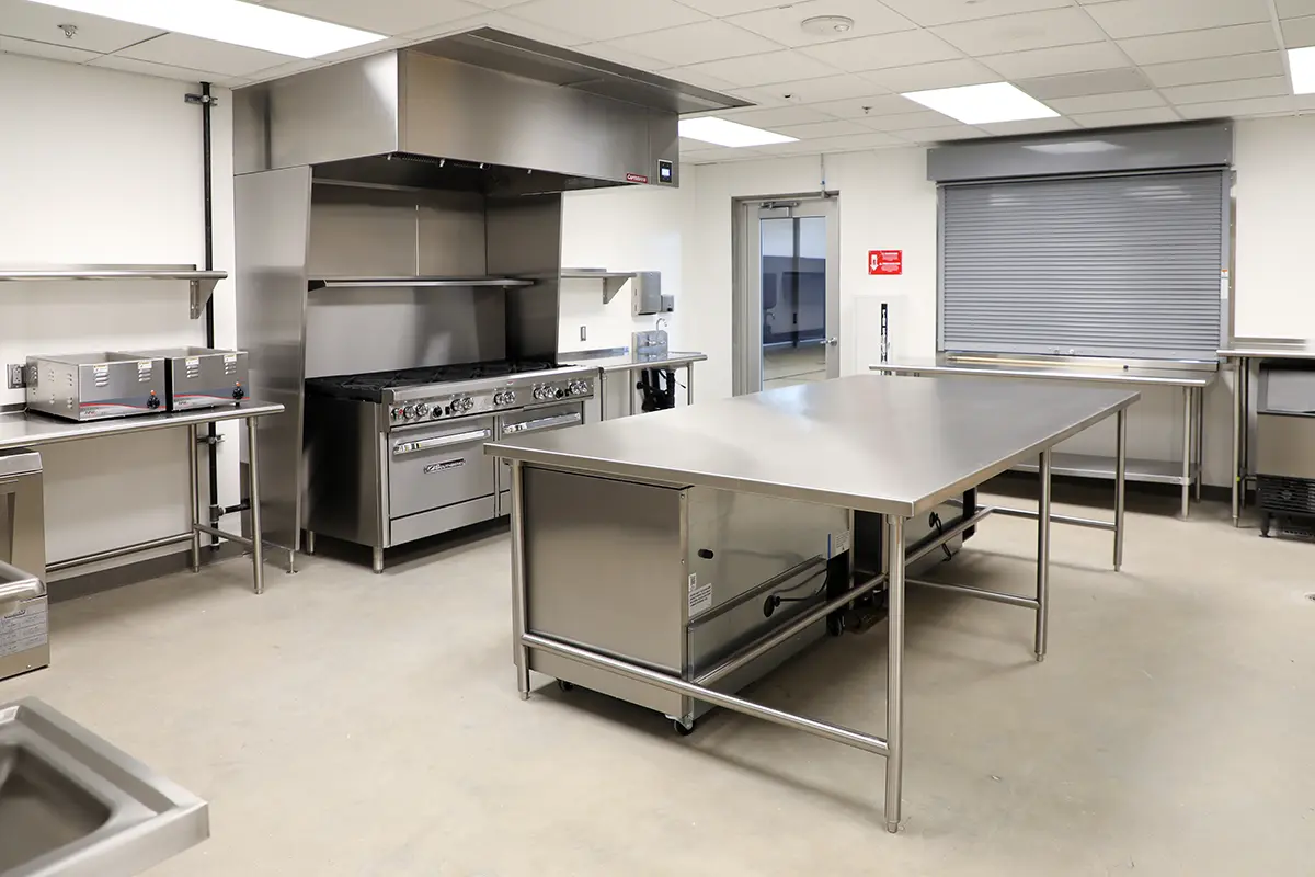 A bright, clean industrial kitchen with stainless steel appliances; a large cooking range, metal countertops, and a prep table, creating a professional ambiance.