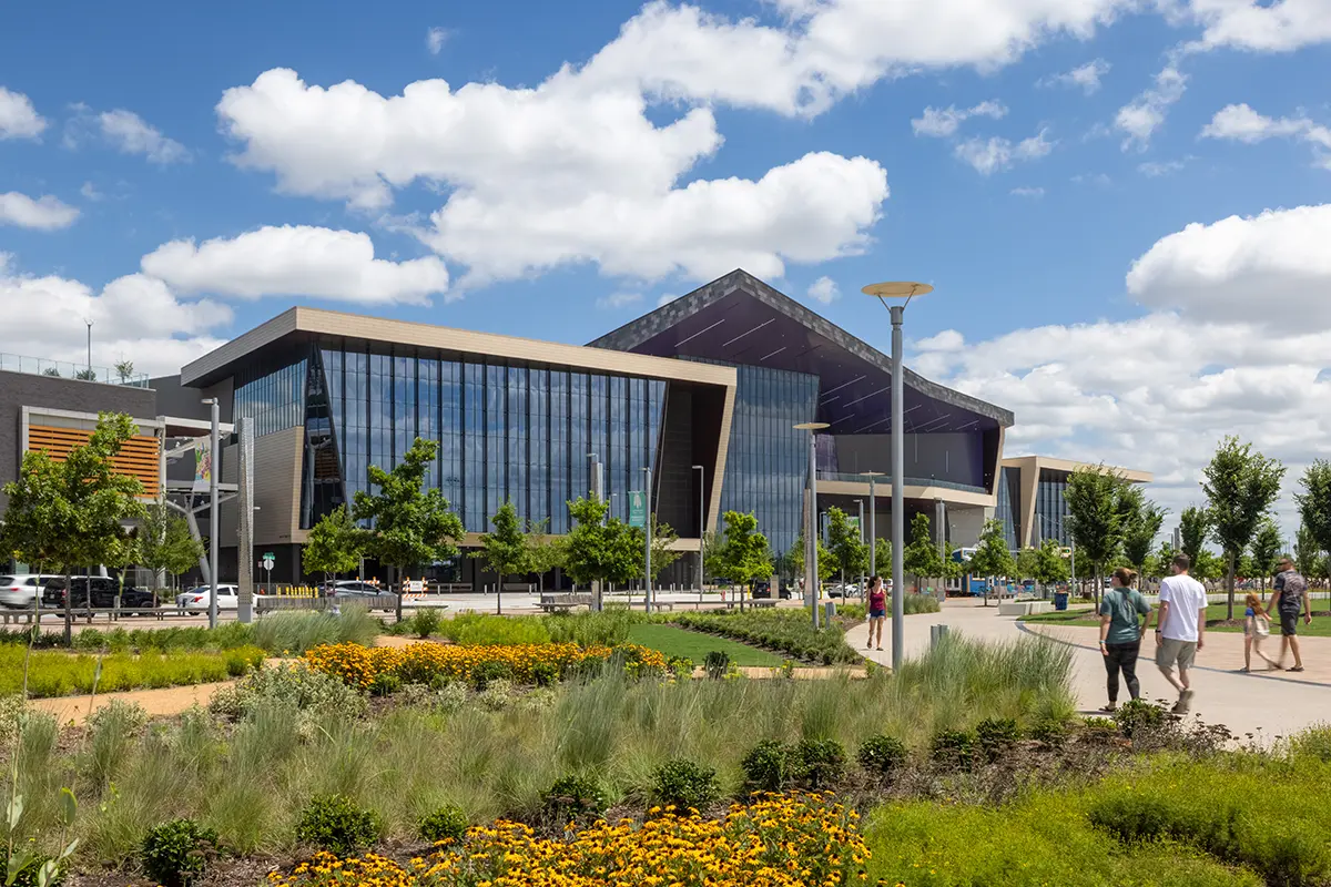 Modern building with glass facade under a bright blue sky and clouds. People stroll along a path surrounded by vibrant gardens and lush greenery.