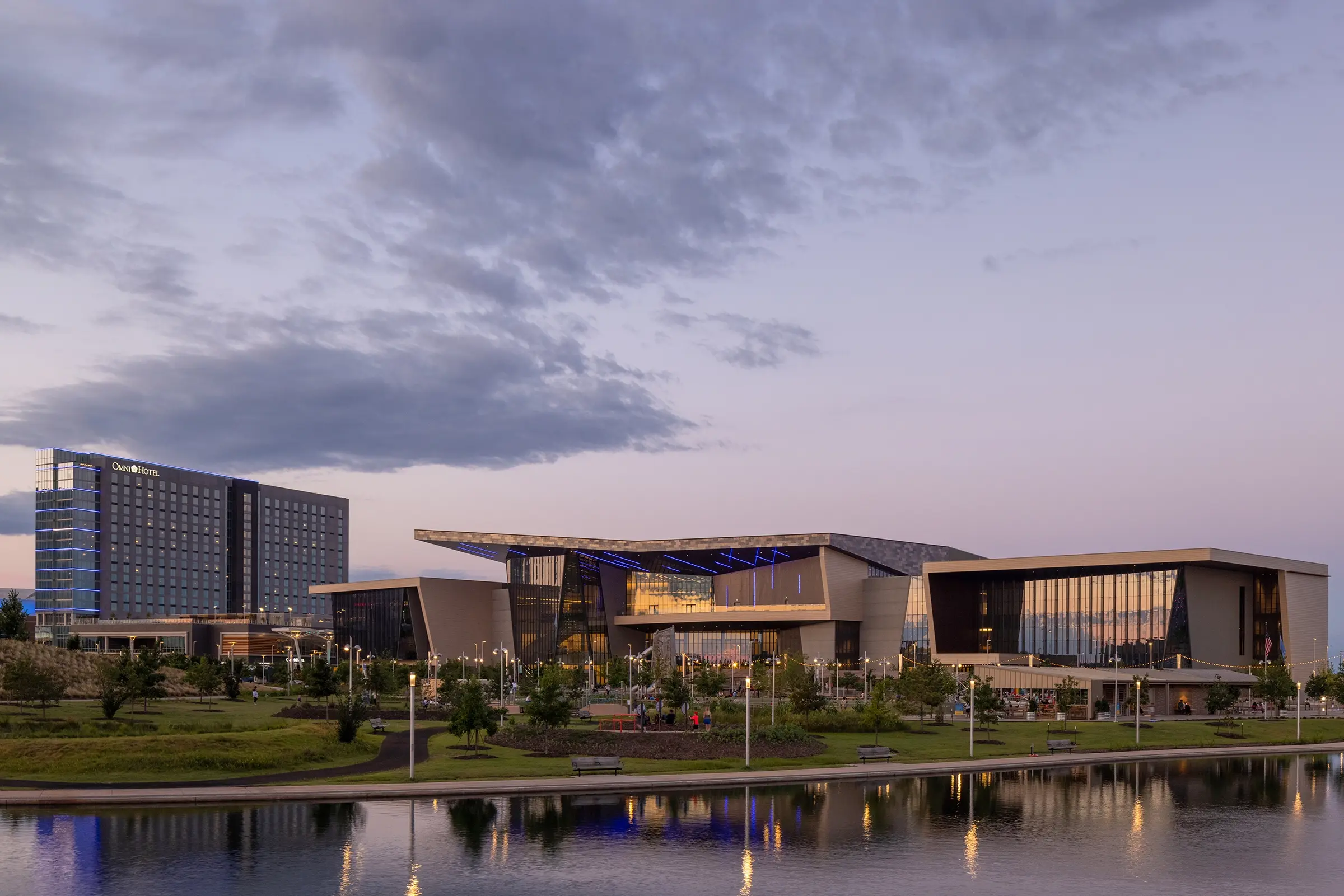 Modern conference center and hotel complex at dusk, with expansive glass facades reflecting soft purple skies. A tranquil pond sits in the foreground.
