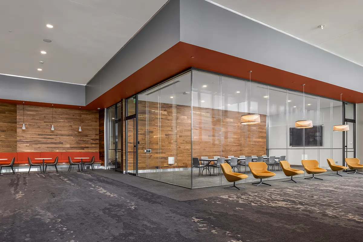 Modern office lobby with glass-walled meeting room, wood paneling, grey and orange tones, pendant lights, and yellow chairs, creating a sleek, inviting atmosphere.