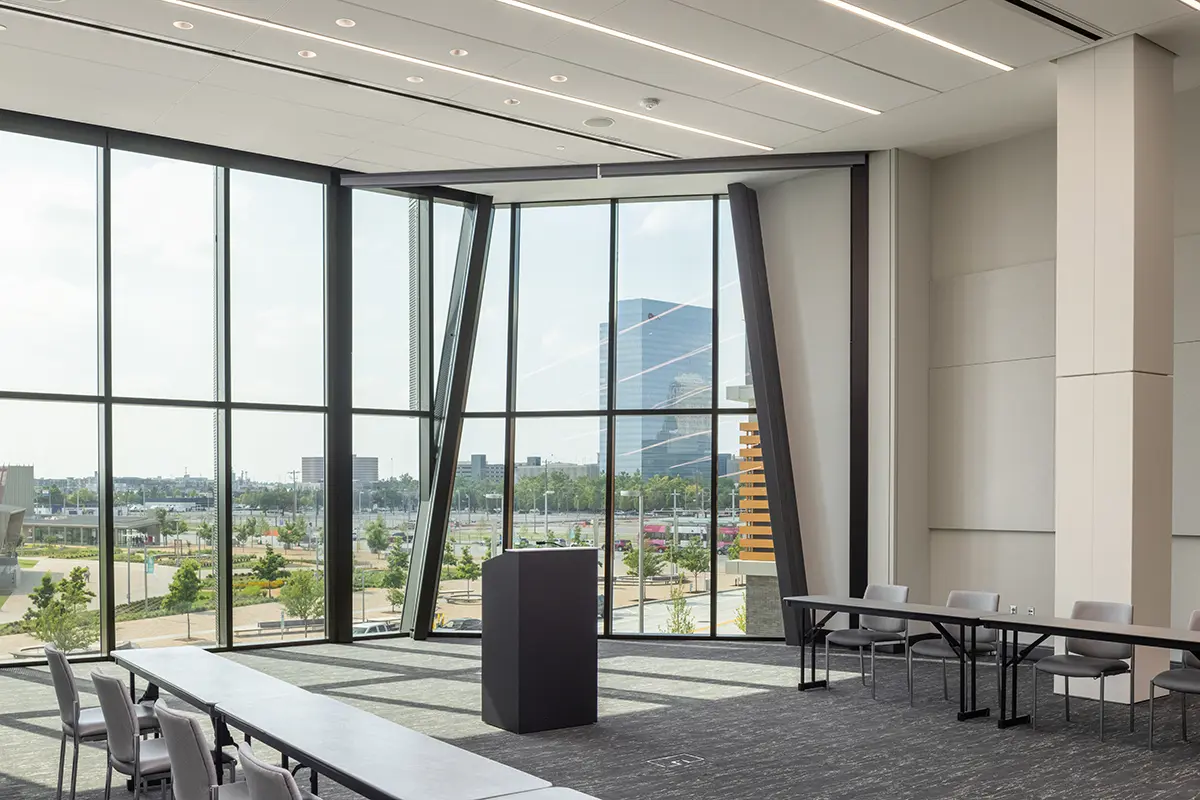 Modern conference room with large angled windows offering a cityscape view. Features include a podium, tables, and chairs, creating a sleek, professional atmosphere.