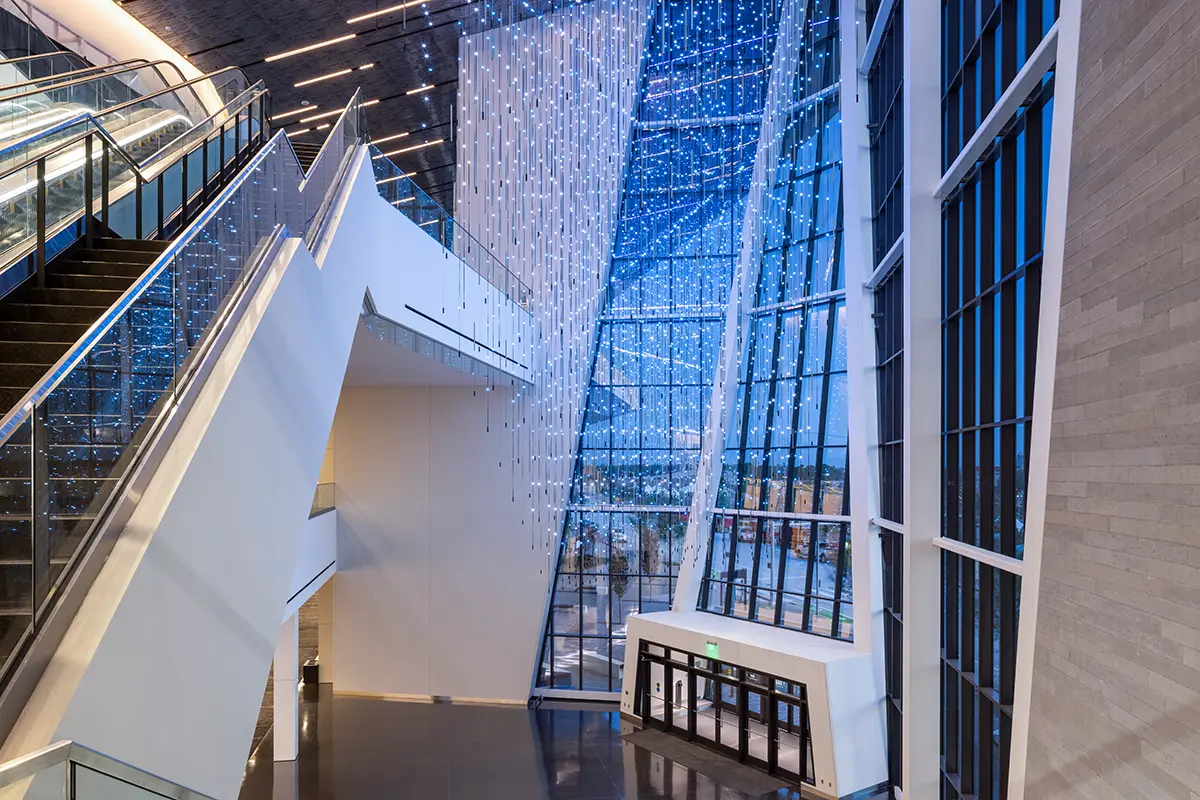 Modern atrium interior with large angled glass walls showcasing a night cityscape, illuminated by cascading ceiling lights, conveying a sleek, futuristic vibe.