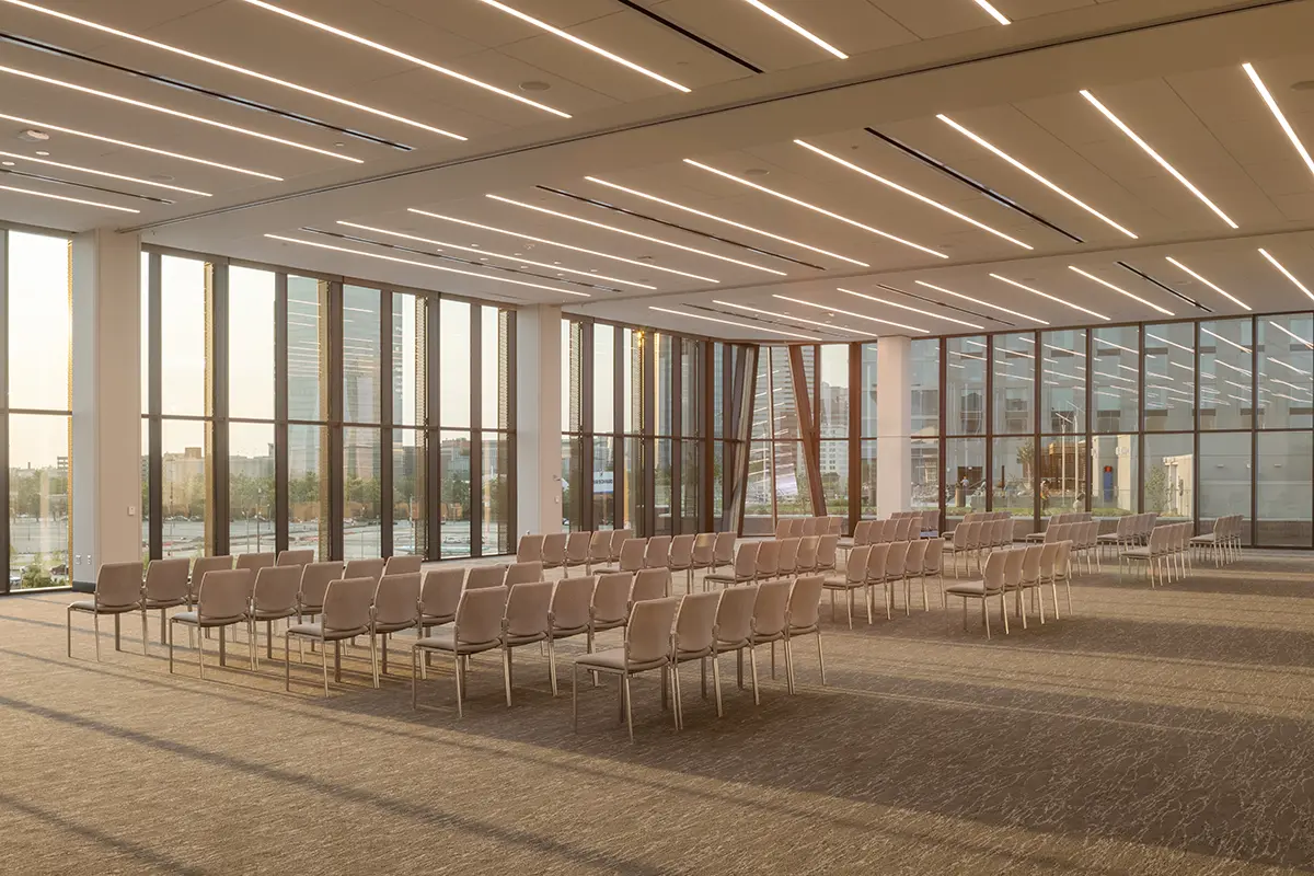 Spacious, modern conference room with rows of empty beige chairs, large windows letting in soft natural light, and sleek linear ceiling lights.