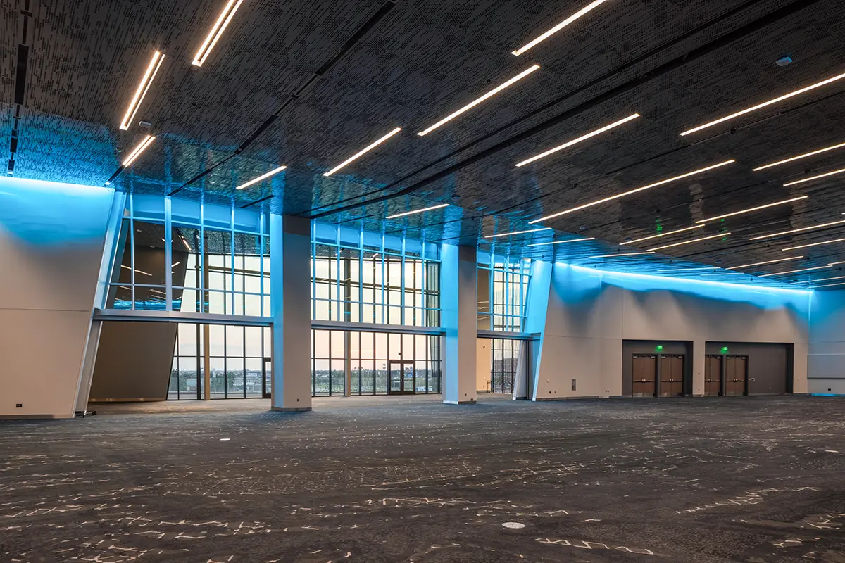 Spacious modern conference hall with high ceilings and sleek lighting. Large windows offer a view of the sunset. Ambient blue light creates a calm atmosphere.