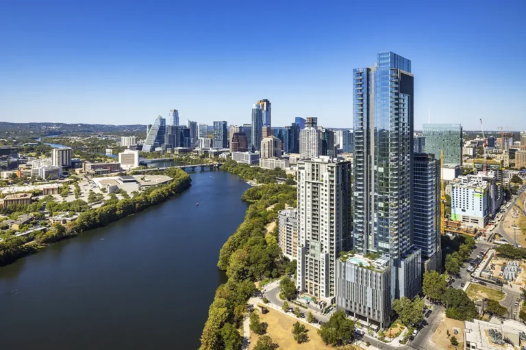 Aerial view of a modern cityscape with a river in the foreground. Tall skyscrapers gleam under a clear blue sky, surrounded by lush greenery.