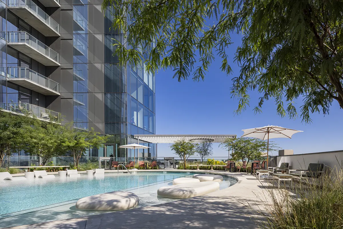 Modern outdoor pool with curved loungers, surrounded by lush greenery and tall glass building. Umbrellas provide shade under a clear blue sky.