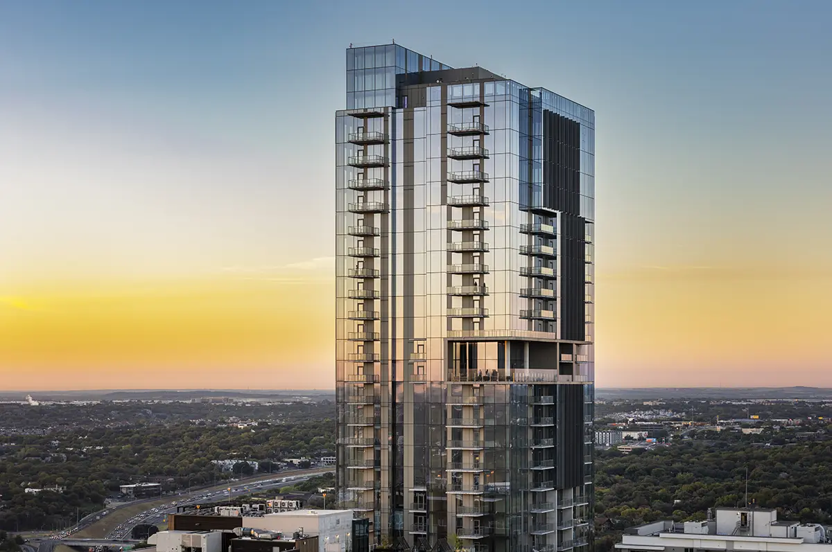 A modern glass skyscraper stands tall against a vibrant sunset sky, reflecting warm hues. The cityscape below features lush greenery and urban sprawl.