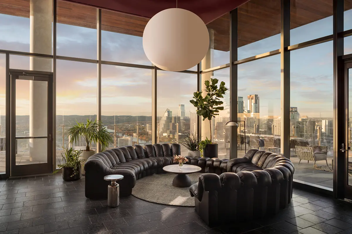 Modern living room with curved leather sofa, round coffee table, large pendant lamp, and plants. Floor-to-ceiling windows showcase a city skyline at sunset.