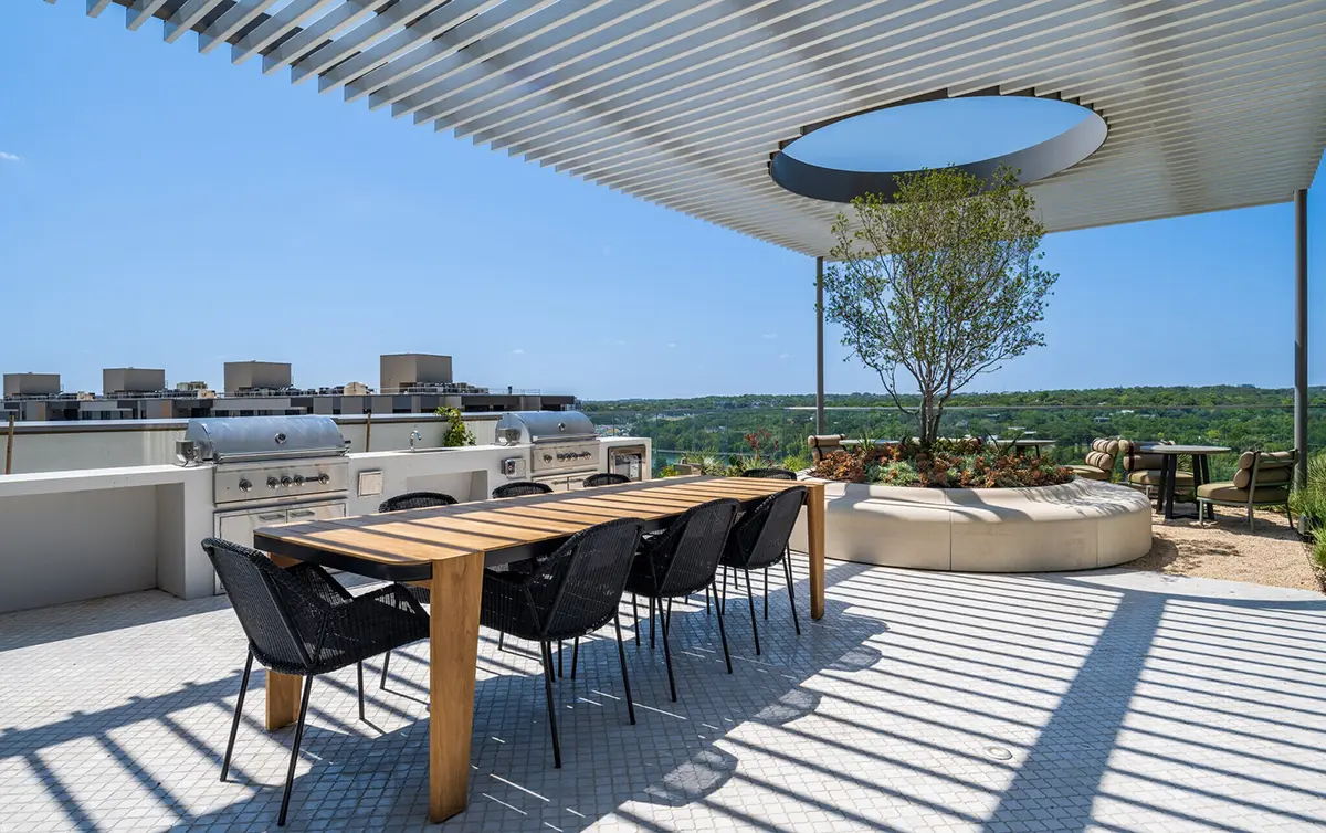 Rooftop patio featuring a long wooden dining table with black chairs, a modern grill, and a circular seating area with a tree. Open pergola casts shadows.