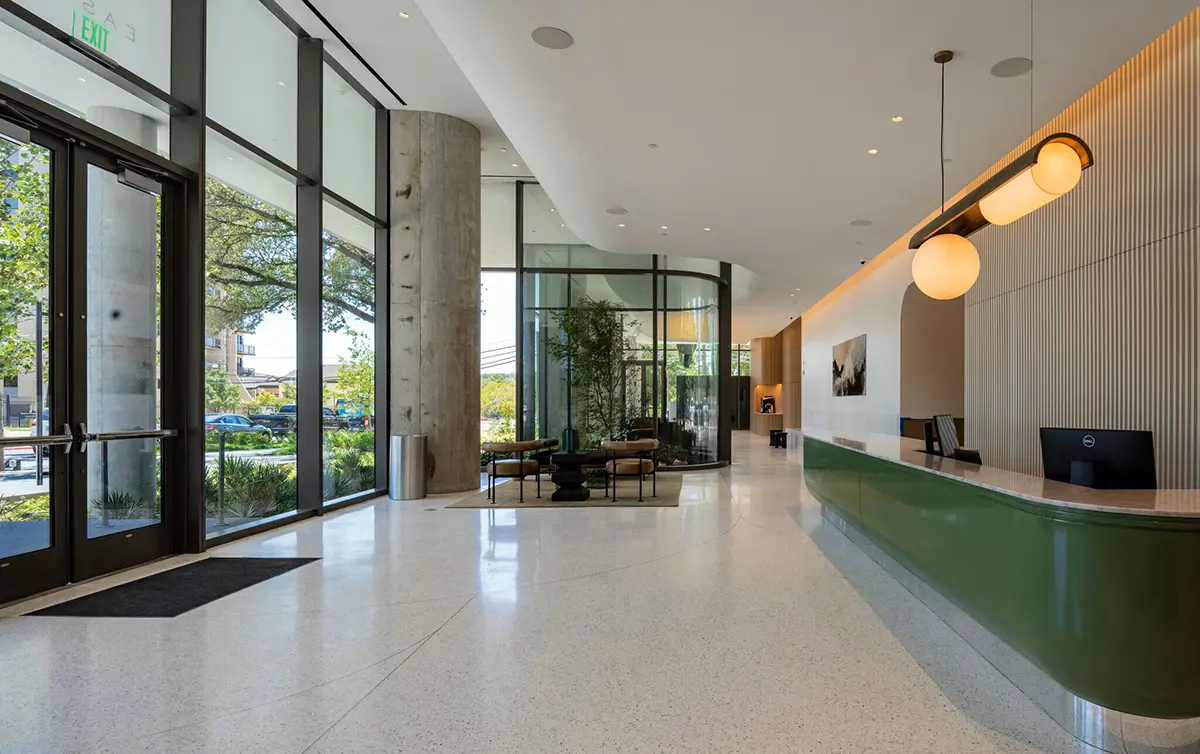 Modern lobby with large glass windows, a sleek green reception desk, and stylish lighting. Natural light highlights the open, airy feel and minimalist decor.