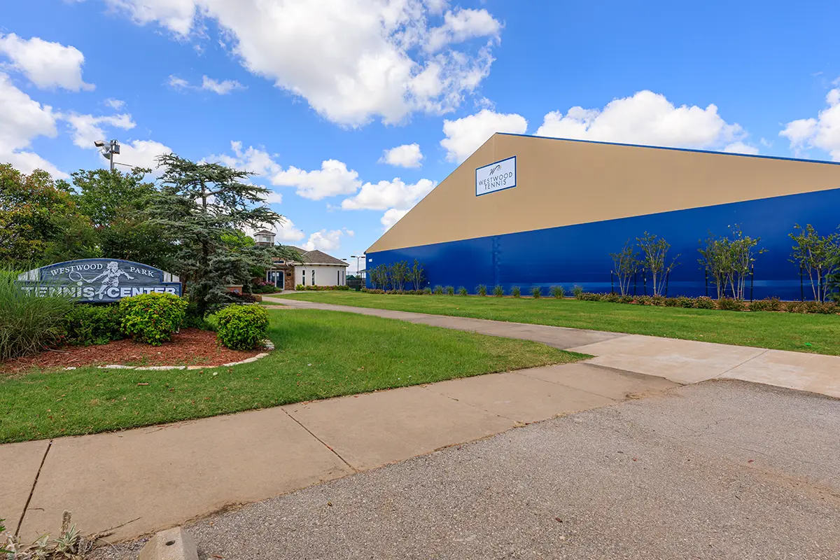 Tennis center with a large blue and beige building, surrounded by green grass and small trees. A sign reads "Westwood Park Tennis Center." Bright day with scattered clouds.