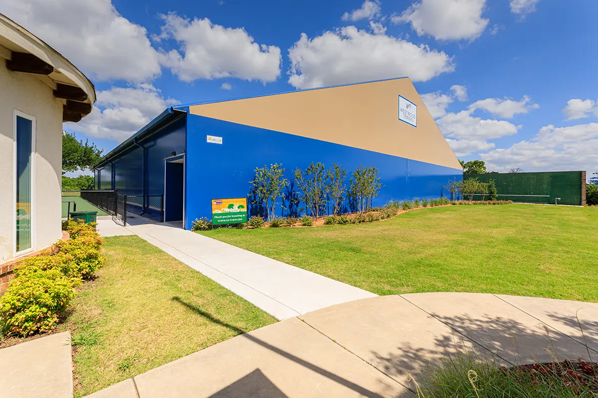 A vibrant blue building with a beige facade stands under a bright, partly cloudy sky. A well-maintained path and green lawn border the structure.