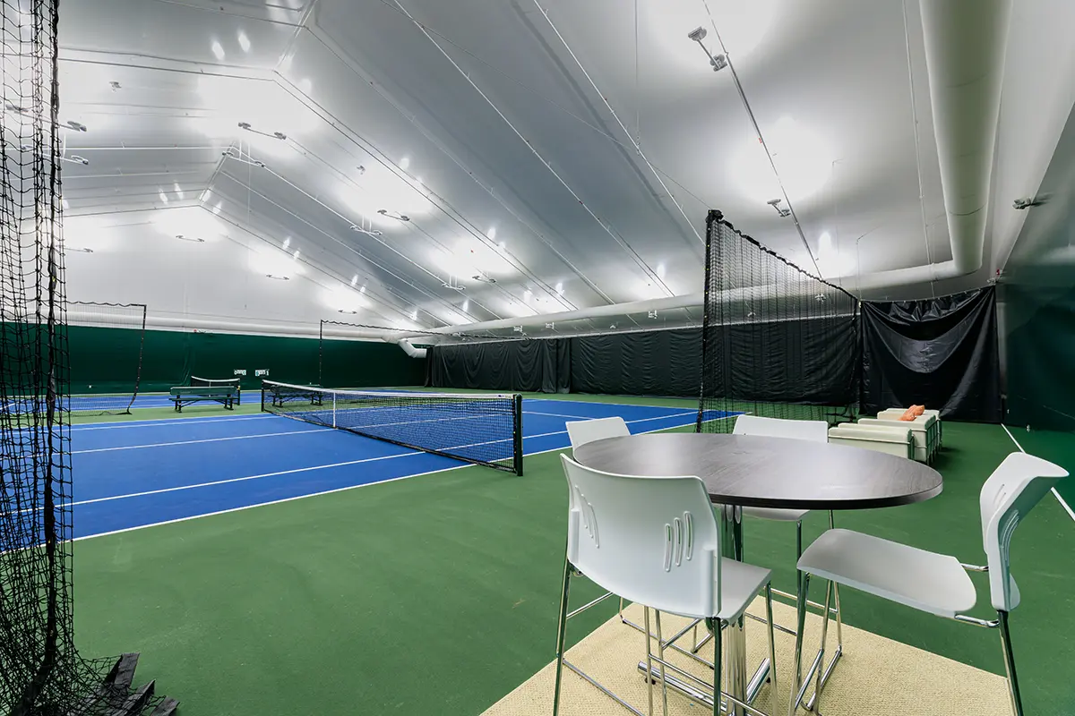 Indoor tennis court with vibrant blue surface and green borders. A table with chairs is in the foreground, creating a relaxed, inviting atmosphere. Bright overhead lights.