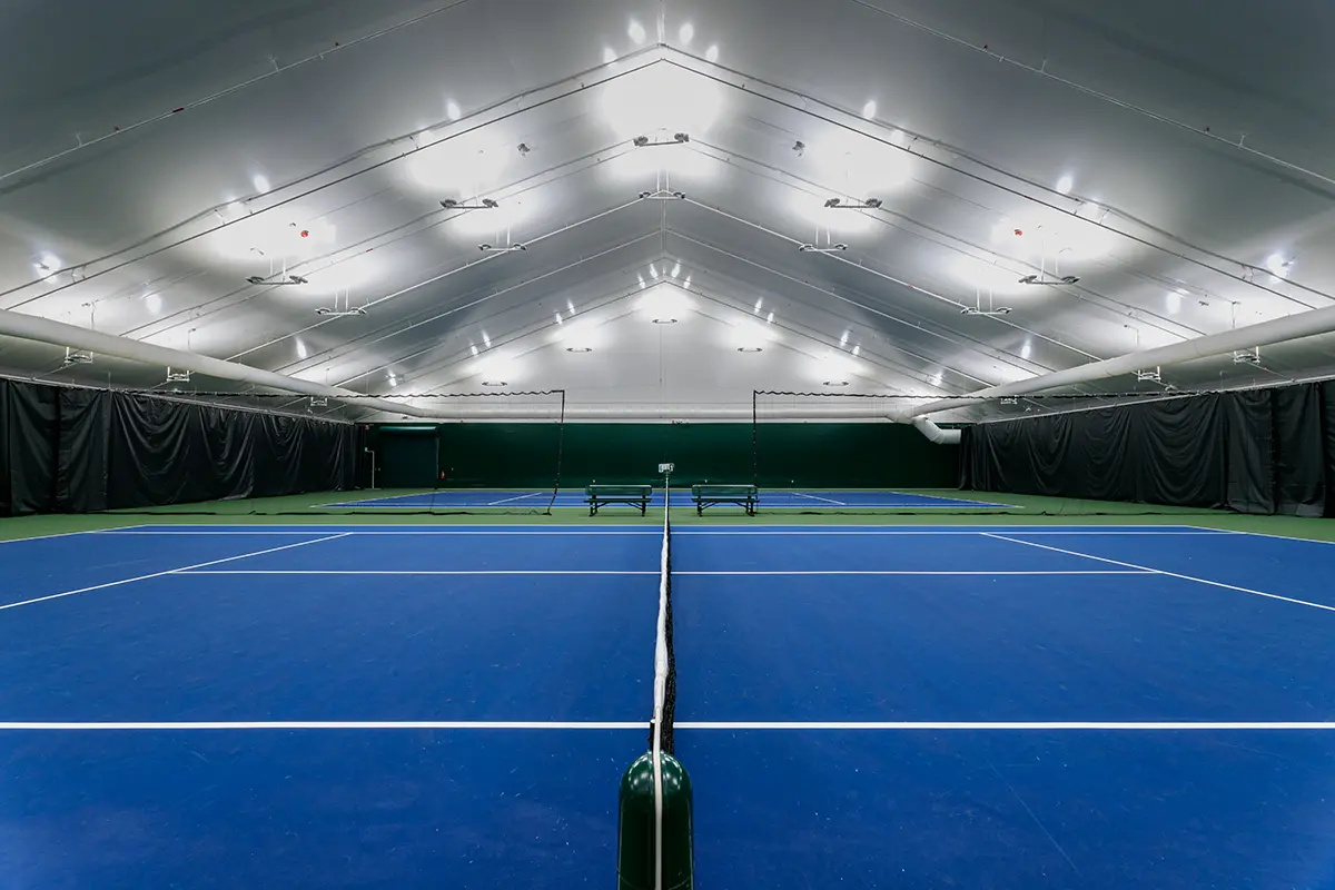 Indoor tennis facility with two blue courts and a high, well-lit ceiling. Black curtains line the walls, creating a clean, modern atmosphere.