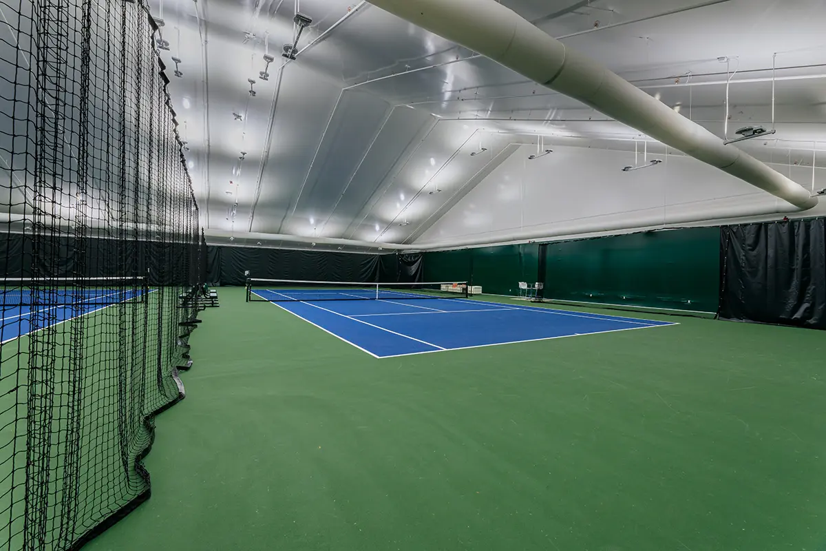 Indoor tennis court with bright lighting, featuring blue playing surfaces and surrounded by a green floor. Black curtains and nets are visible, creating a clean, modern atmosphere.