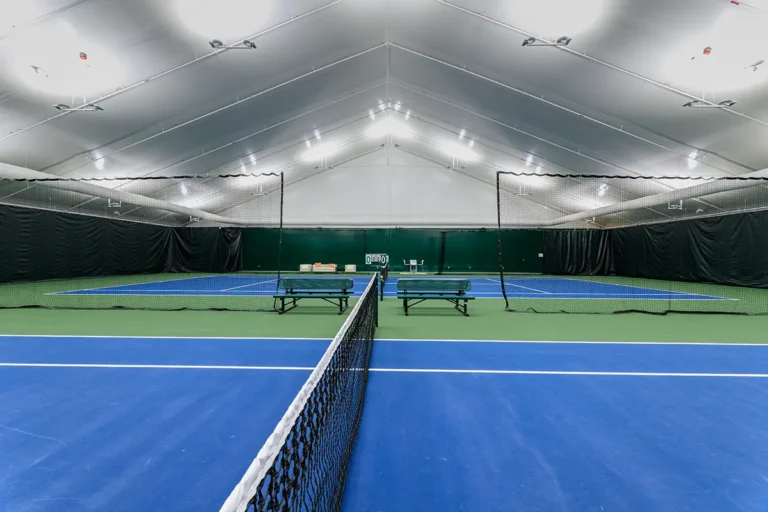 Indoor tennis court with blue and green flooring under a tall, white canopy ceiling. Rows of lights above illuminate the net and empty benches.