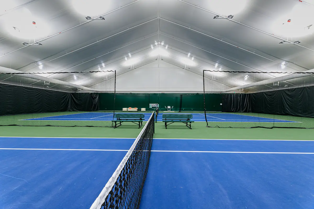 Indoor tennis court with blue and green flooring under a tall, white canopy ceiling. Rows of lights above illuminate the net and empty benches.