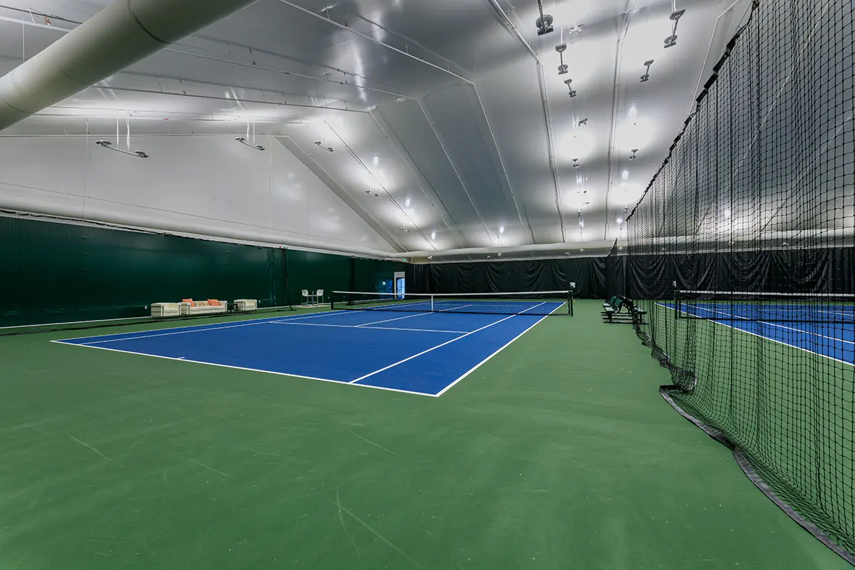 Indoor tennis court with blue and green surfaces, white lines, and a high, arched ceiling. Bright lighting creates a clean, spacious atmosphere.