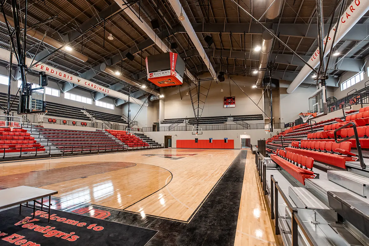 Spacious indoor basketball court with polished wood floors, surrounded by red and black bleachers. Bright lighting gives an inviting, energetic tone.