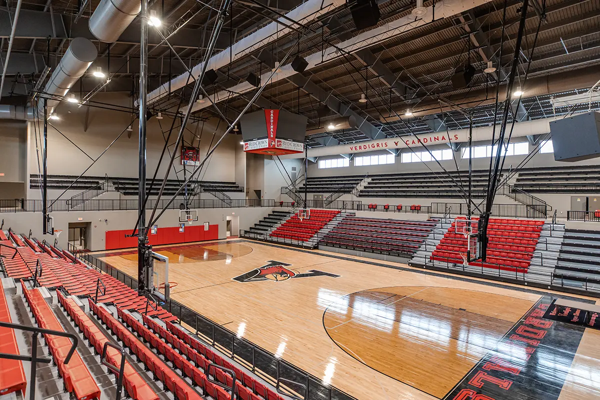 Spacious indoor basketball court with polished wooden floors and red bleachers. Overhead lighting highlights the team's logo at center court.