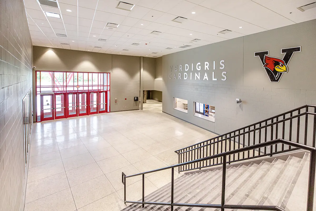 Bright school lobby with red-framed glass doors, gray walls, and tiled floor. A mural reads "VERDIGRIS CARDINALS" beside a red cardinal logo.