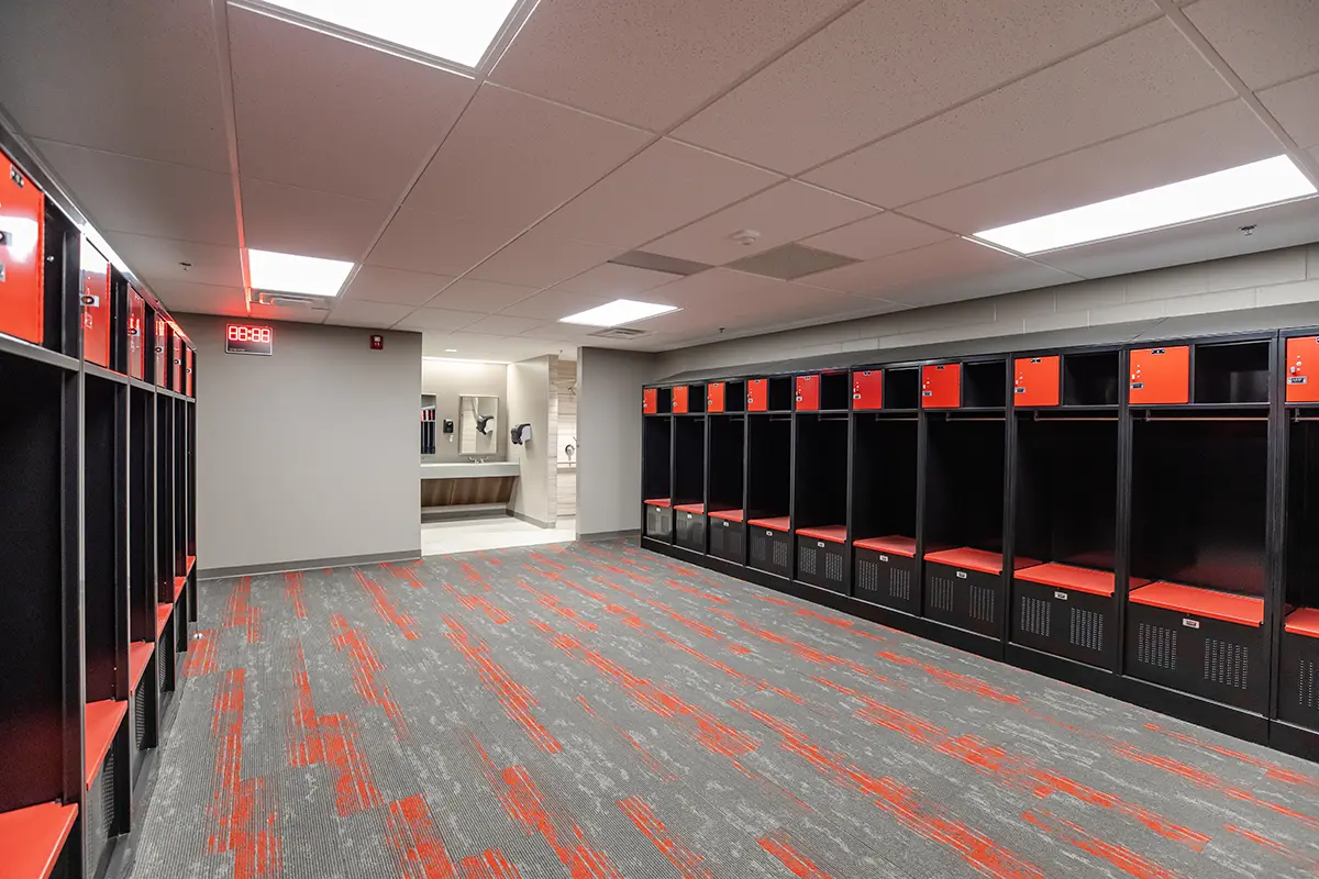 Modern locker room with black and red lockers lining the walls, red-accented gray carpet, and bright overhead lighting, leading into a bathroom area.