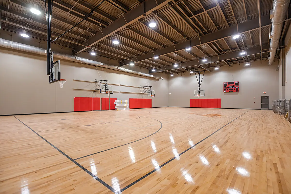Spacious indoor basketball court with polished wooden floors, bright lighting, red wall padding, and multiple hoops, conveying an inviting sports atmosphere.