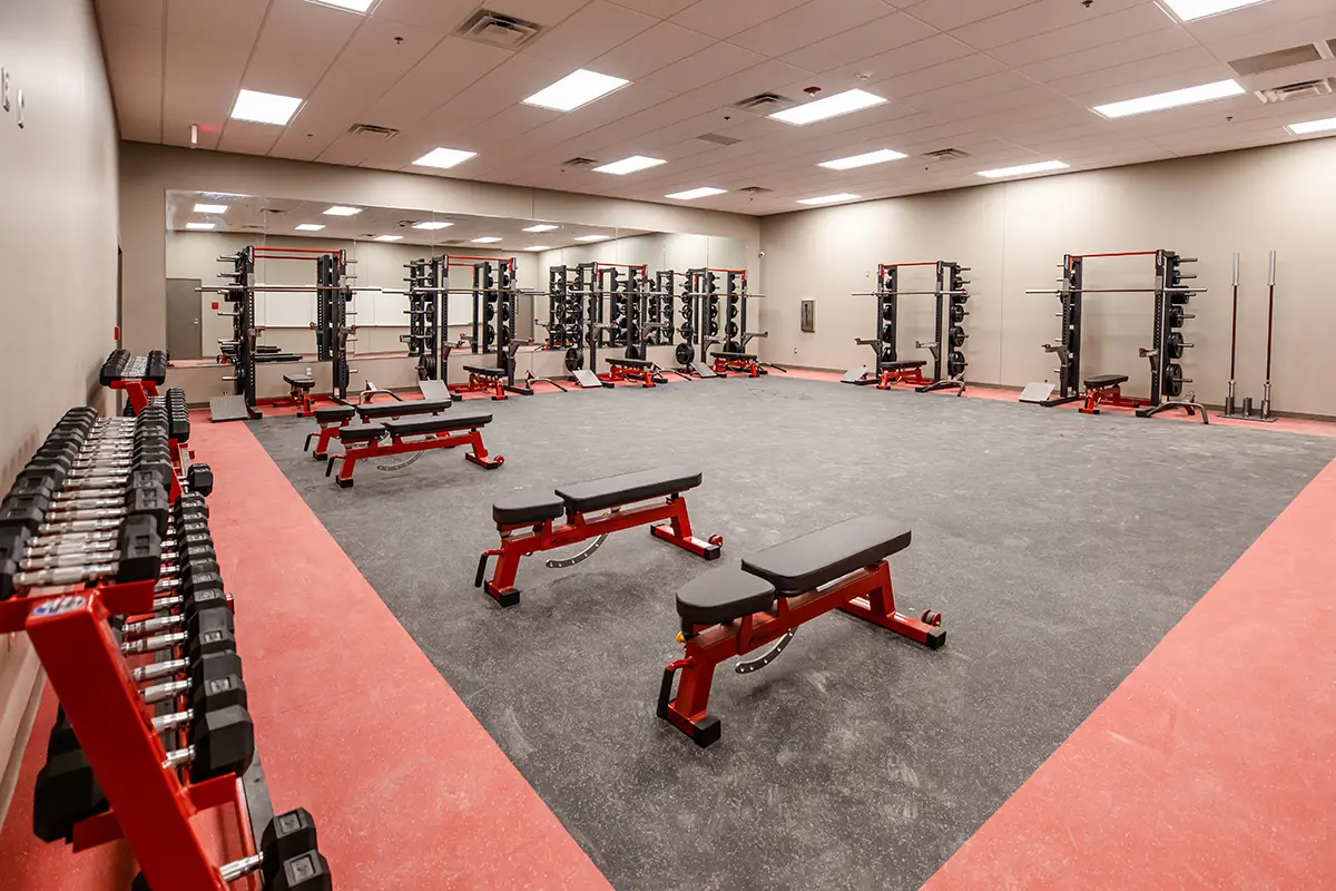 Spacious weight room with red benches, dumbbells, and racks of weights. Bright lighting and mirrored wall create a clean, professional gym vibe.