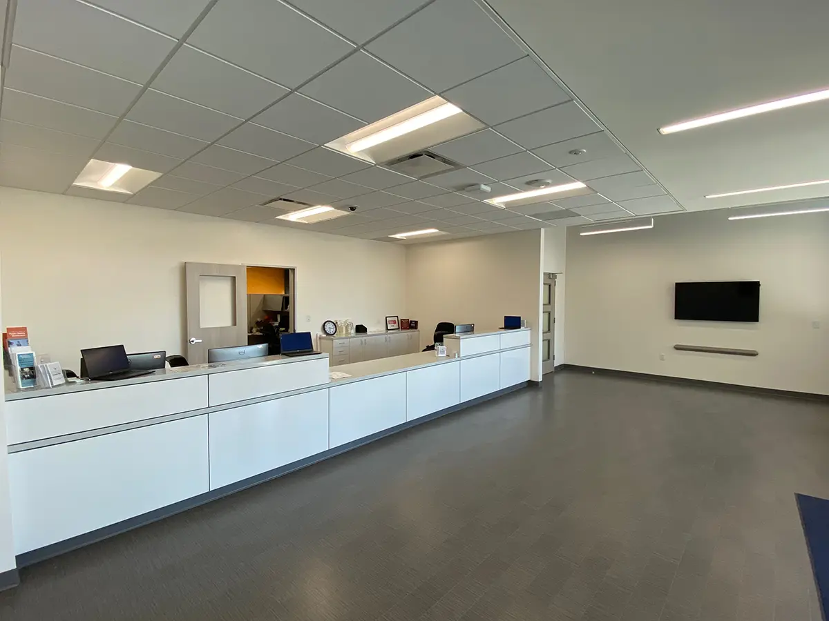 Wide-angle view of a modern, minimalist office reception area with a long white desk, computers, and office supplies. Walls and ceiling are off-white, creating a bright, professional atmosphere.